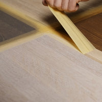 Eine Hand zieht Abklebeband zwischen unterschiedlich gefärbten Holzmusterflächen ab.