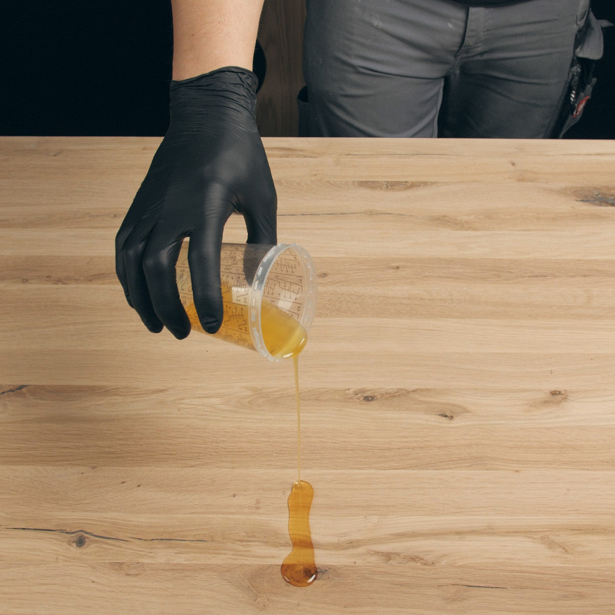 Eine behandschuhte Hand gießt das Öl-Härter-Gemisch aus einem Messbecher auf eine Holzplatte